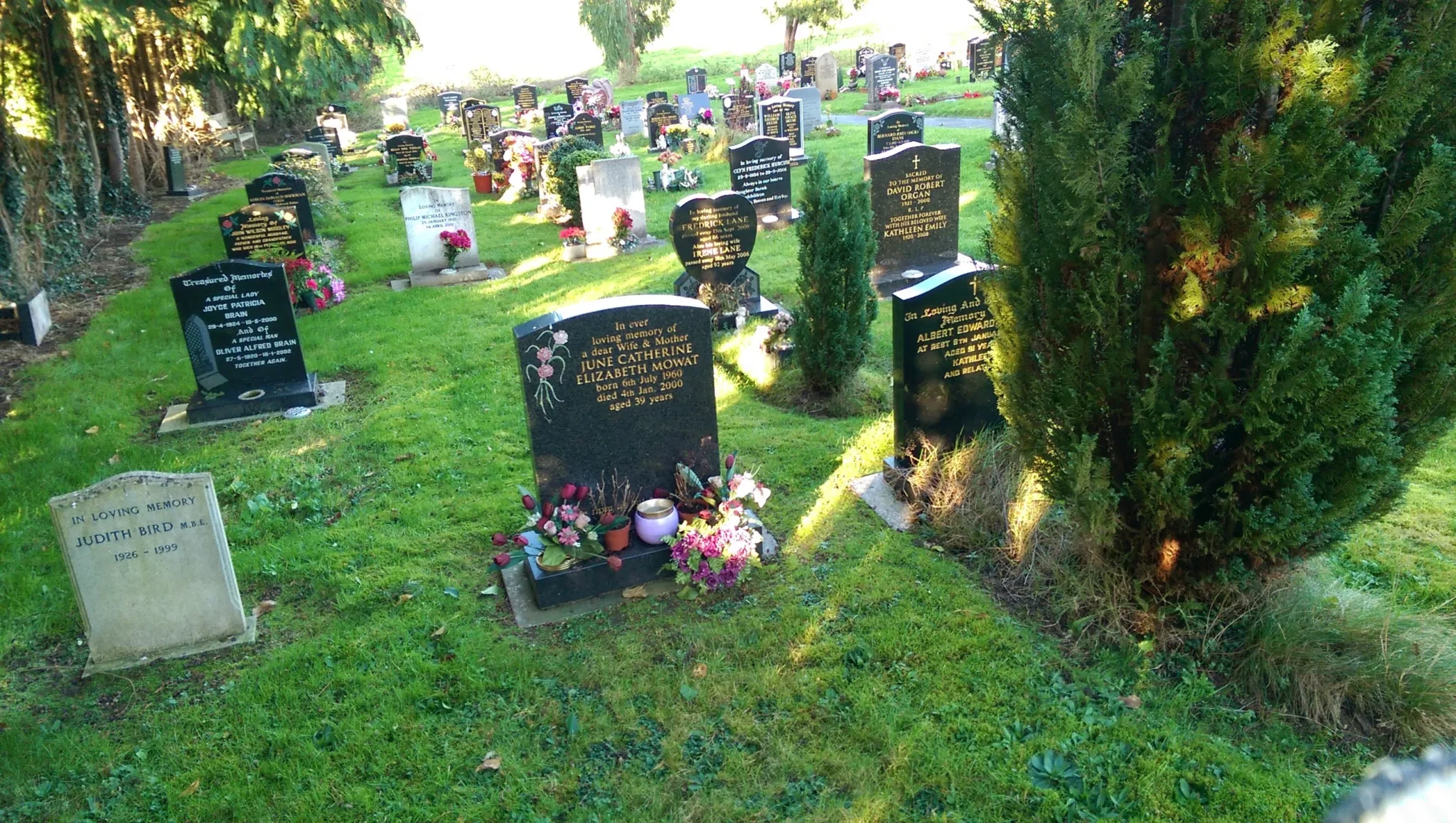 A cemetery with several headstones crafted by Memorial Masons, some adorned with flowers and wreaths, surrounded by green grass and trees on a sunny day.