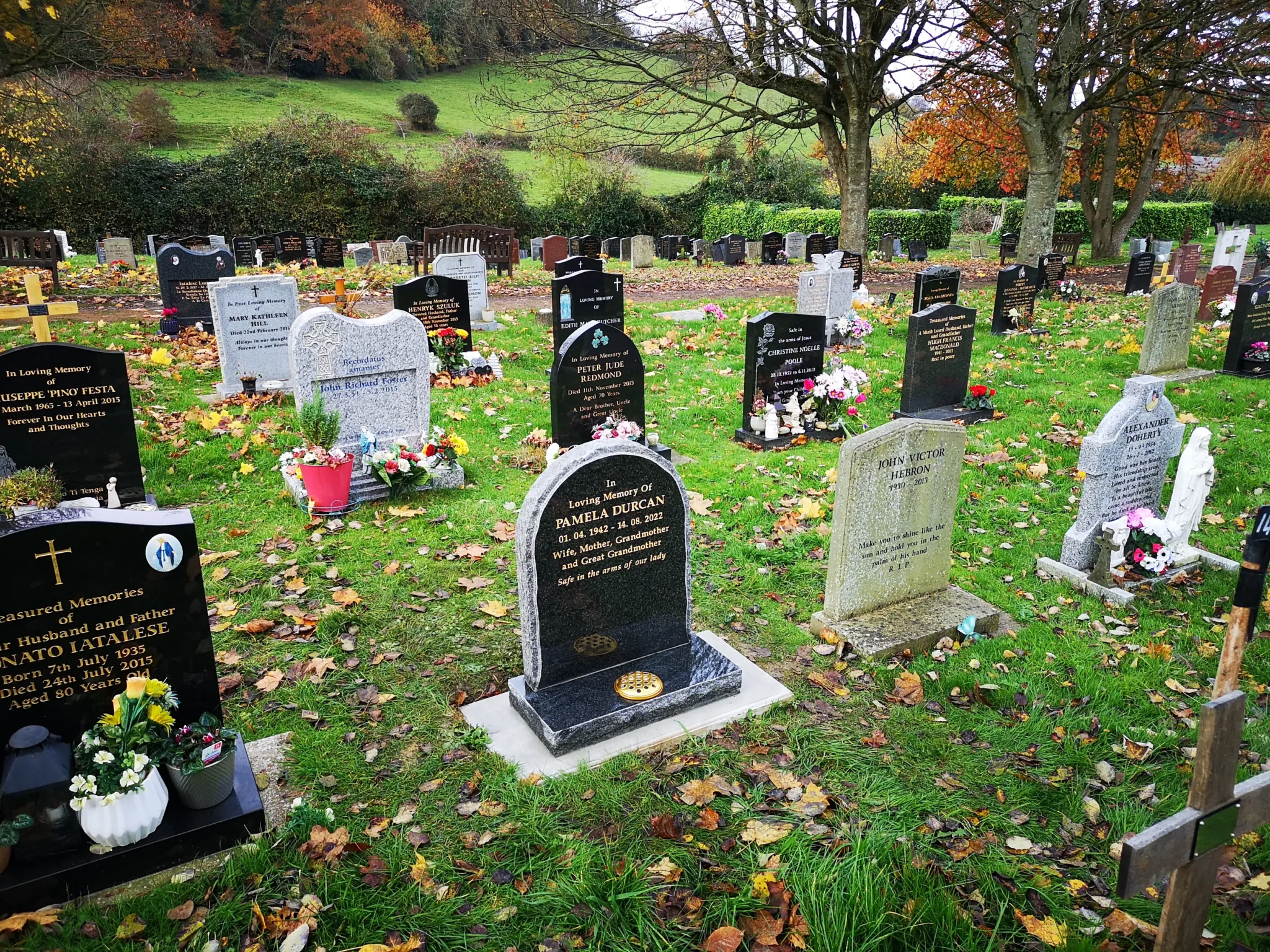 A cemetery with numerous headstones and memorials, some decorated with flowers and wreaths. Fallen autumn leaves cover the grass, and trees with colorful foliage line the background under a cloudy sky, cared for by skilled Memorial Masons.