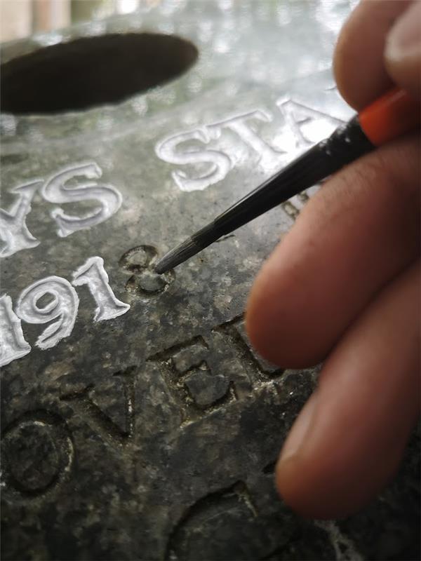A hand uses a small paintbrush to carefully apply white paint to engraved letters and numbers on a dark stone surface, focusing on the number "1918," a delicate process often seen in Stone Restorations for headstones and cremation memorials.
