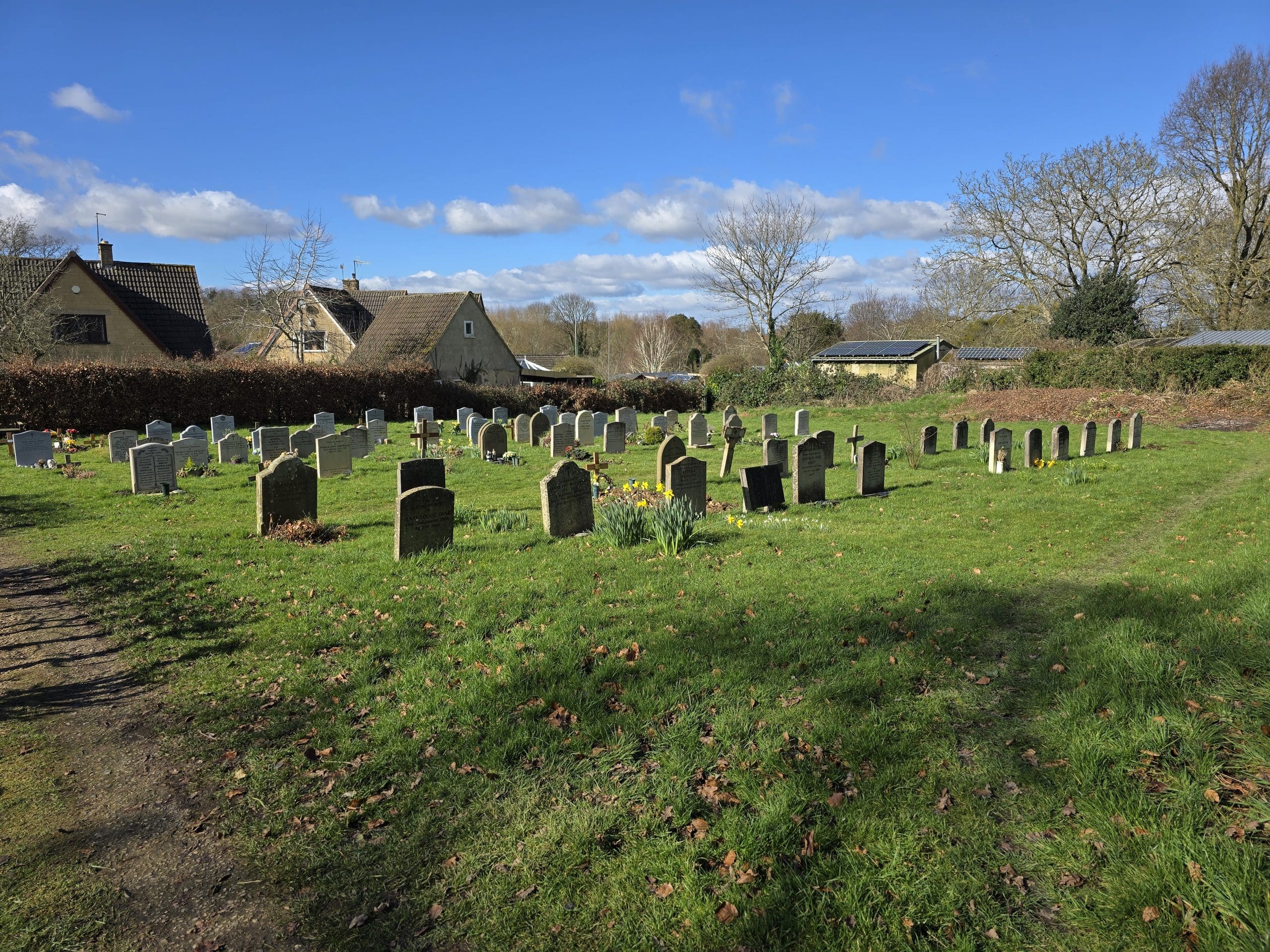 A peaceful cemetery with rows of headstones stands on green grass under a blue sky with scattered clouds. Leafless trees and a few houses are visible in the background, reflecting the work of skilled Memorial Masons.