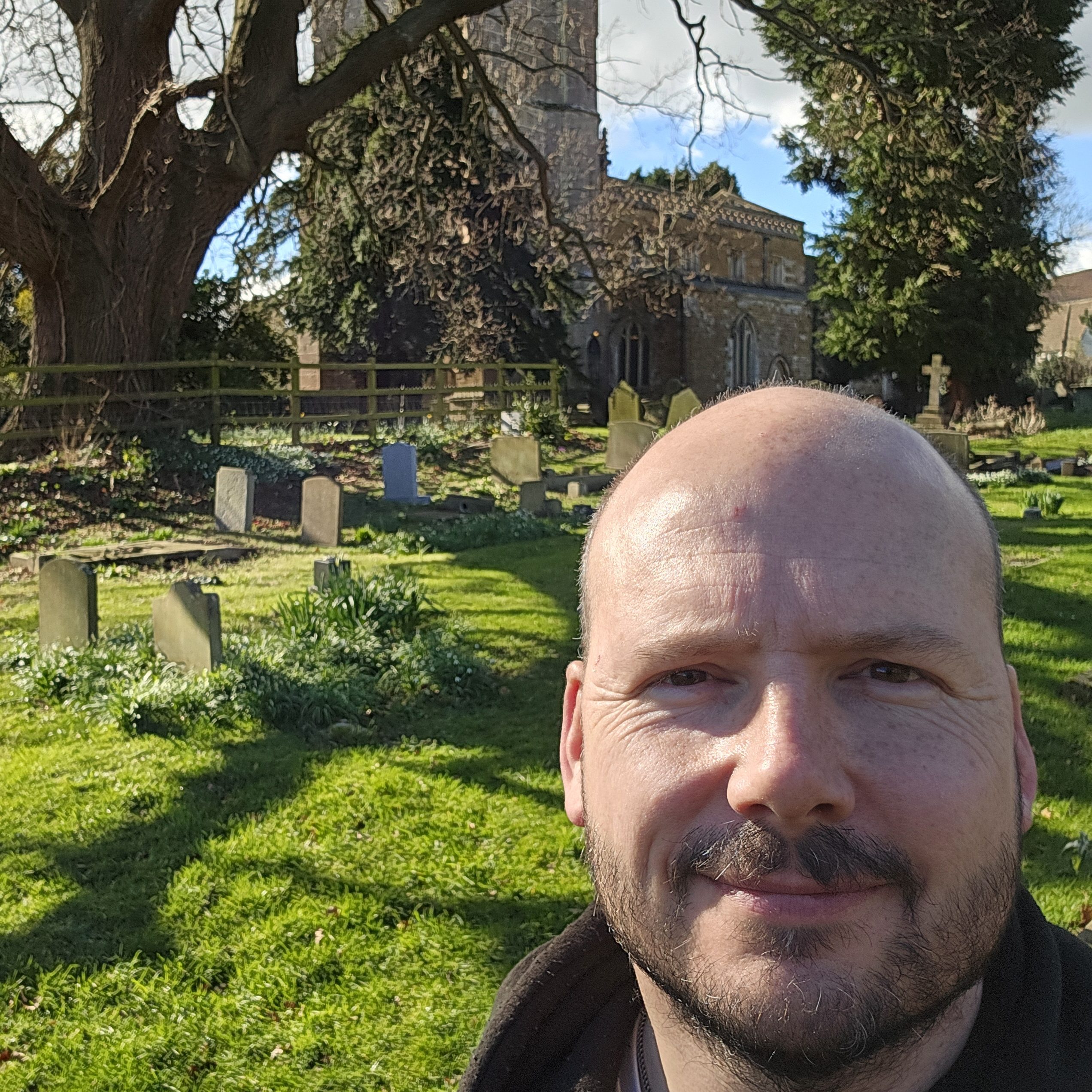 A man with a beard and mustache takes a selfie in a sunny graveyard, surrounded by old headstones, green grass, trees, and a stone church—capturing the quiet beauty of expertly crafted memorial masons’ work.