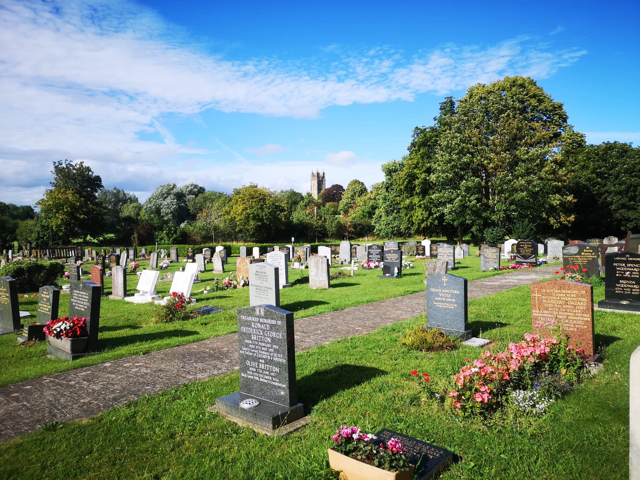 A cemetery with rows of headstones and flower arrangements on a bright, sunny day. Green grass and trees surround the area, with a church tower visible in the distance under a blue sky with scattered clouds, crafted by Memorial Masons.