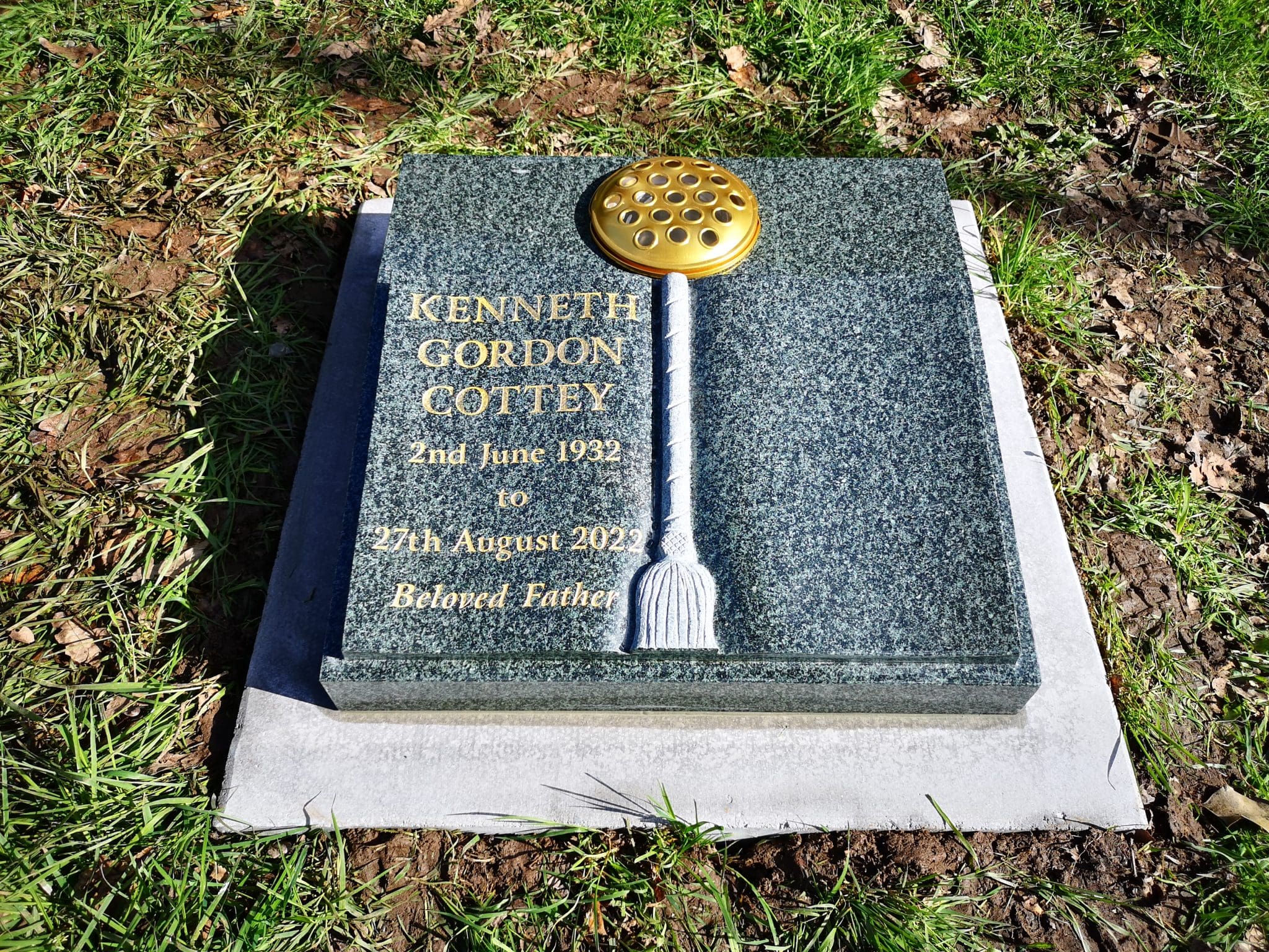 A gravestone by Memorial Masons for Kenneth Gordon Cottey (1932–2022), with gold lettering and a brass flower holder on top, set on grass with fallen leaves. The inscription reads: "Beloved Father.