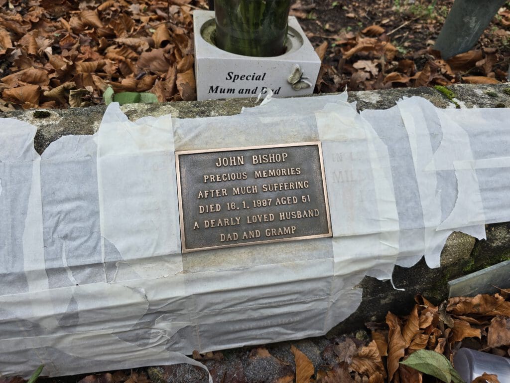 A memorial plaque on a stone, surrounded by fallen leaves, reads: "John Bishop, precious memories after much suffering, died 18.1.1987 aged 51, a dearly loved husband, dad and gramp." A floral tribute is on top, labeled "Special Mum and Dad.
