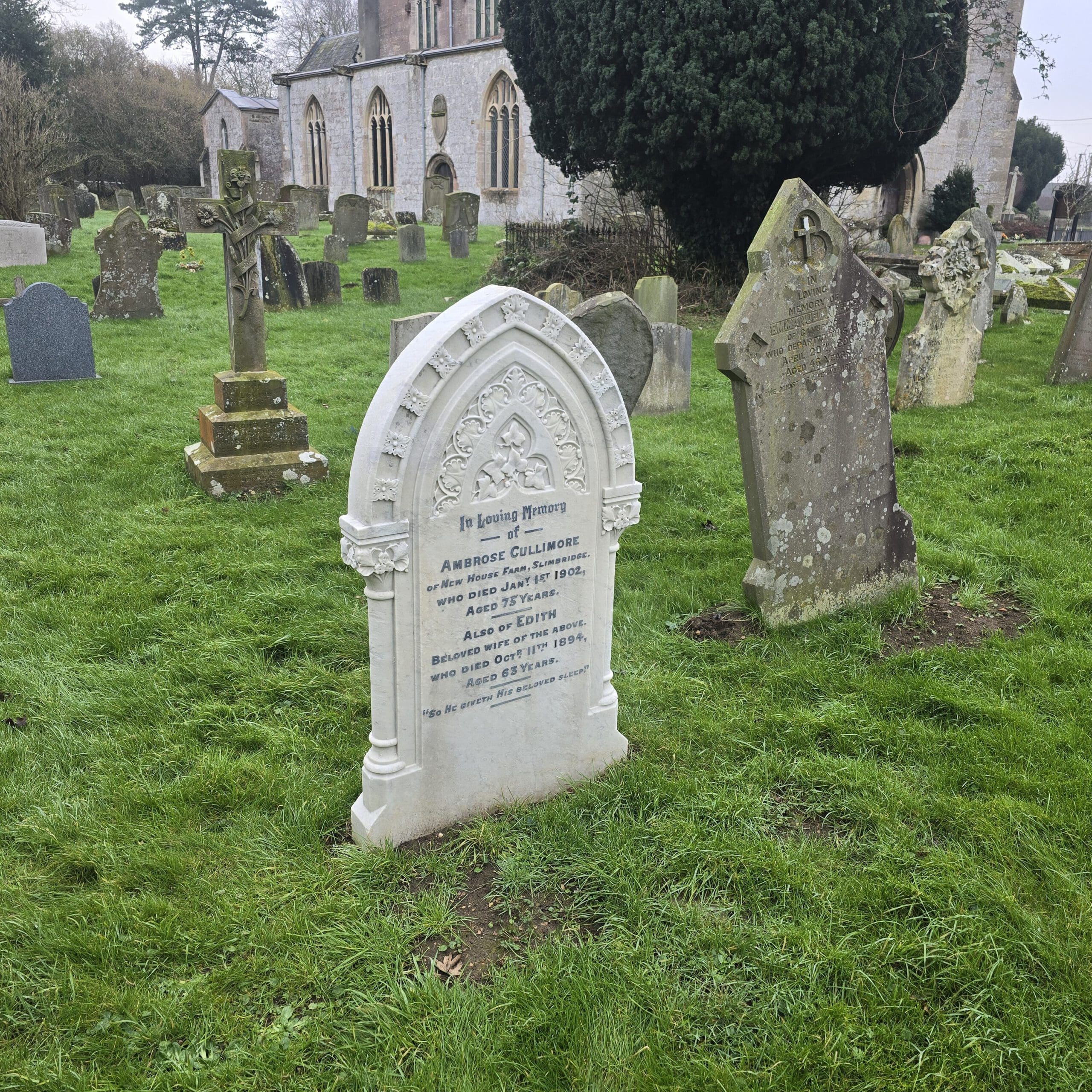 A historical cemetery with several weathered tombstones on green grass. One prominent white tombstone has visible engraved text. In the background, a large stone building, possibly a church, is partially visible with trees surrounding it.