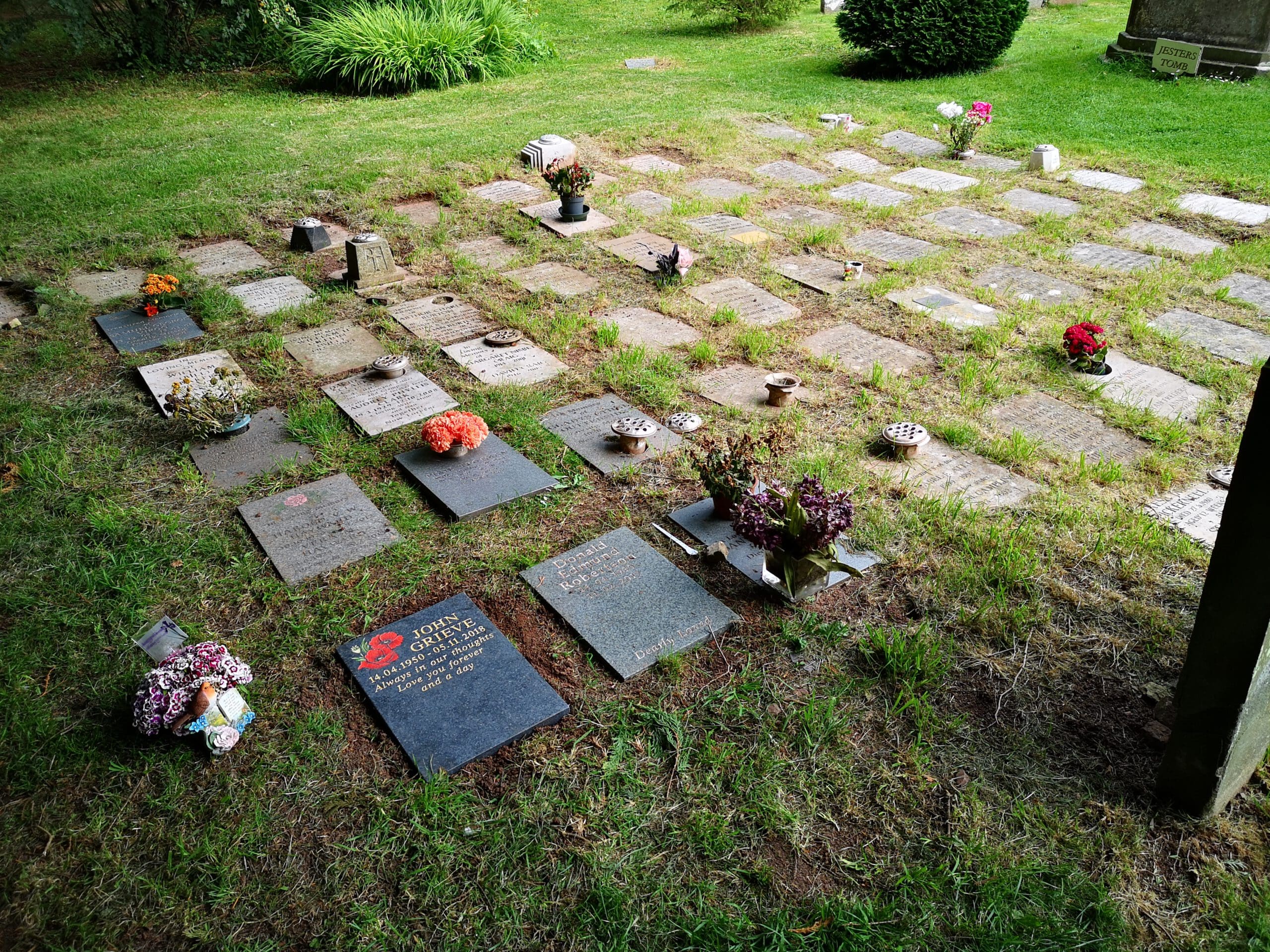 A grassy cemetery area with rows of flat headstones. Some headstones have flowers in pots or small arrangements placed on top. The grass and nearby bushes are lush and green, indicating a well-maintained area.