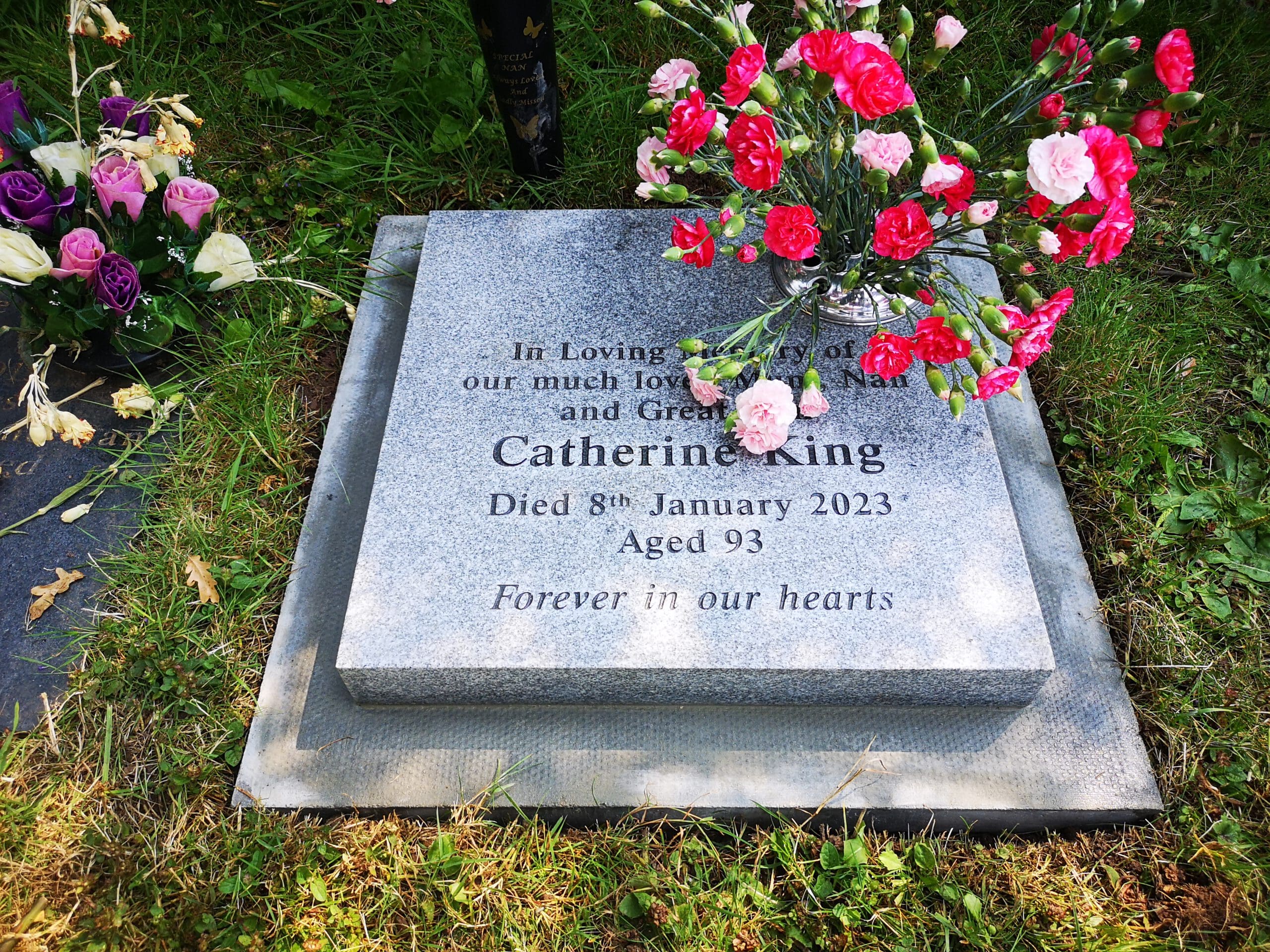 A gravestone in a grassy area with flowers on top. The inscription reads: "In Loving Memory of our much loved Mum, Nan, and Great Nan, Catherine King. Died 8th January 2023 Aged 93. Forever in our hearts." Colorful flowers are laid around it.